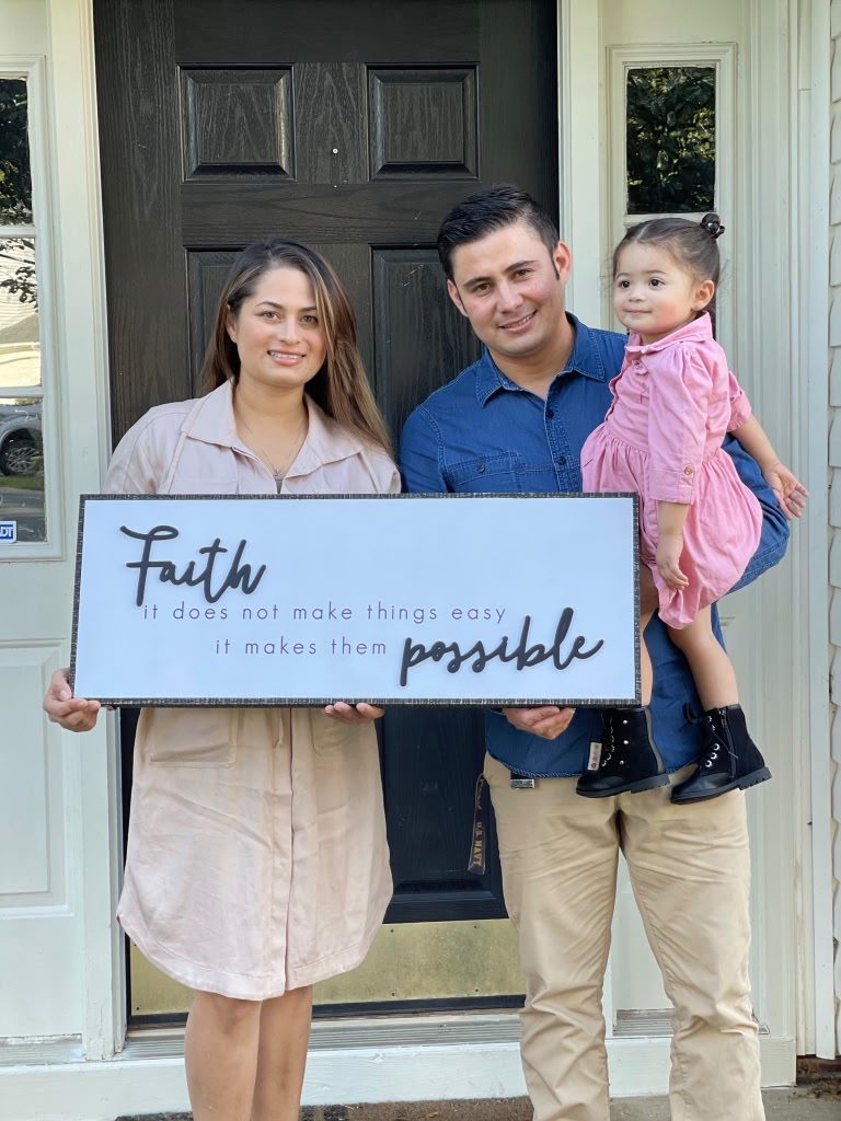Couple with their toddler holding a 'Faith makes things possible' sign at the door of their new home