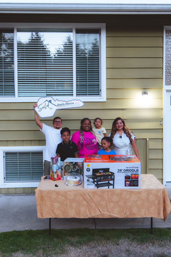 A family standing beside a table of housewarming gifts in front of their new home