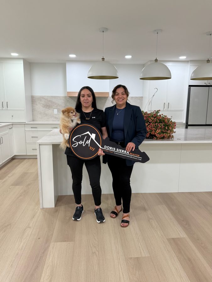 Two women and their small dog holding a Sold by Doris Sierra key sign in the kitchen of their new home